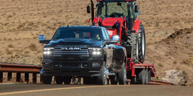 A black RAM dually truck towing a large red tractor on a trailer along a desert highway.
