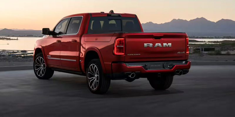Rear-three-quarter view of a cherry red Ram 1500 Tungsten pickup truck parked at sunset, overlooking a body of water and distant mountains.