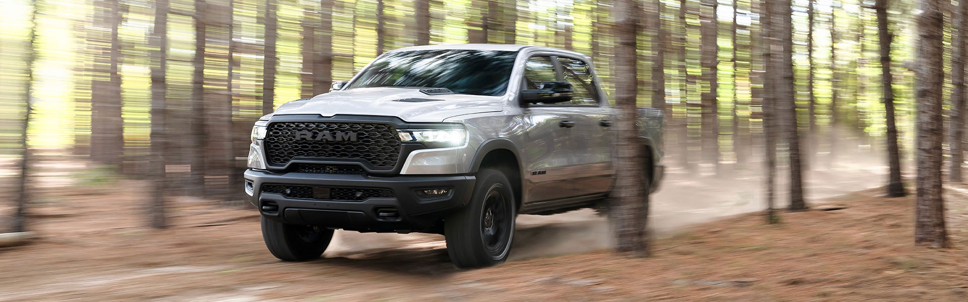 A silver Ram 1500 RHO pickup truck driving through a forest trail, kicking up dust.